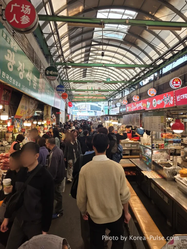 A crowded indoor alleyway of Gwangjang Market in Seoul, filled with locals and tourists walking under a high arched glass roof. Traditional food stalls with colorful signs selling mung bean pancakes (Bindaetteok) and beef tartare (Yukhoe) line both sides of the vibrant market street.