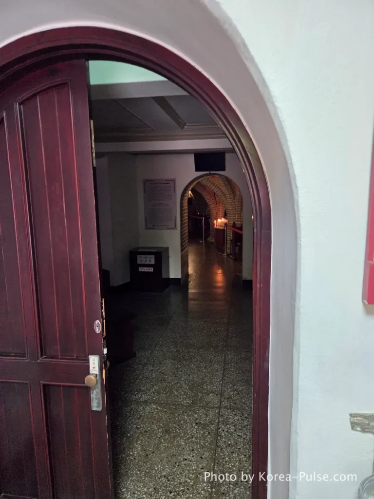Myeongdong Cathedral: Seoul's Gothic Soul, Hidden Above the Shopping District 16 Wooden arched doorway leading into the dimly lit Crypt Chapel beneath Myeongdong Cathedral — underground sanctuary with relics of Korean martyrs