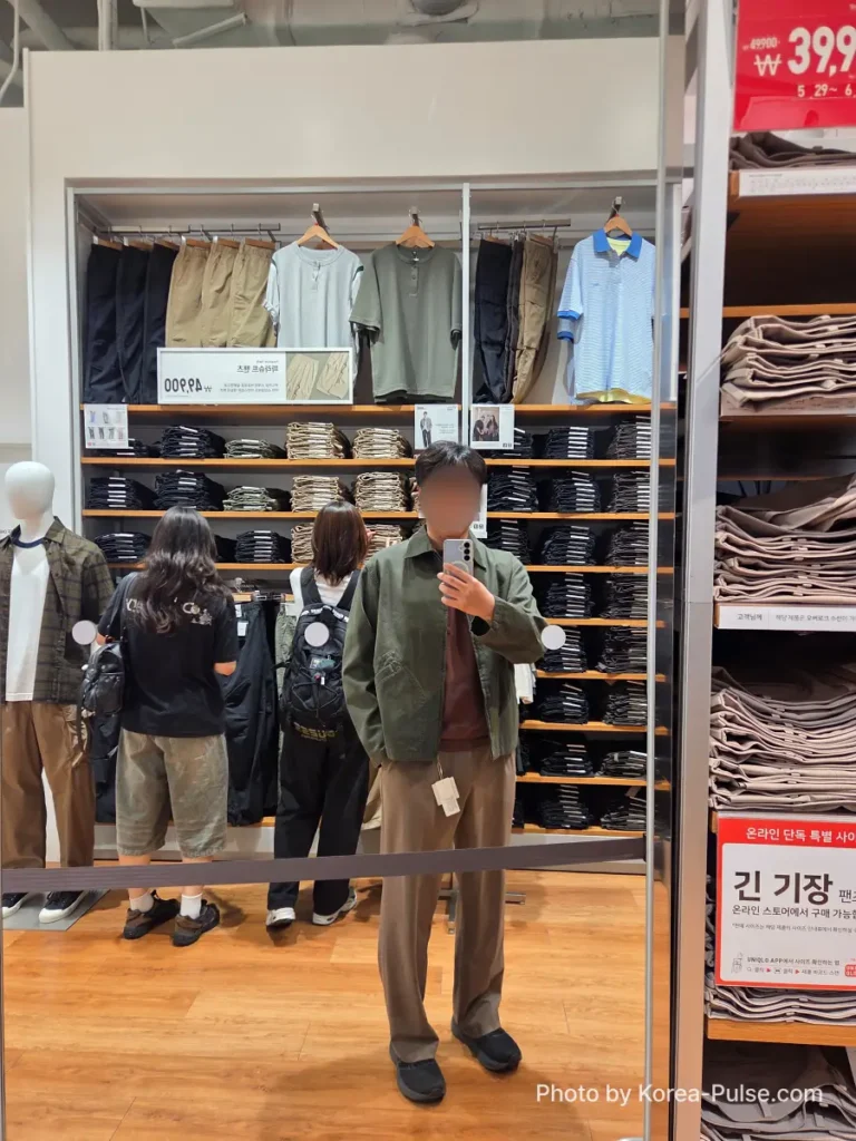 Mirror selfie of a man trying on the olive green Uniqlo JW Anderson Zip-up Blouson inside a Uniqlo Seoul store, with other shoppers and clothing racks visible in the reflection
