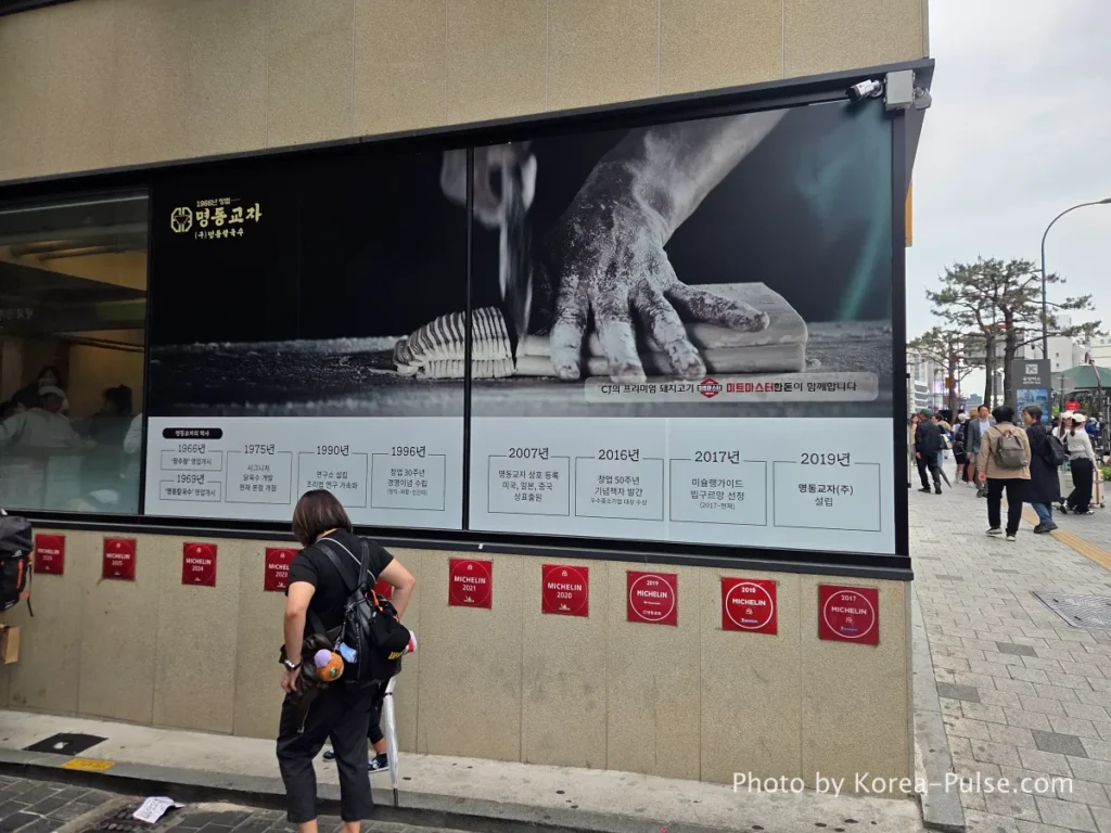 Side wall of Myeongdong Kyoja restaurant displaying full history timeline from 1966 to present alongside row of annual Michelin Bib Gourmand plaques [Myeongdong street]