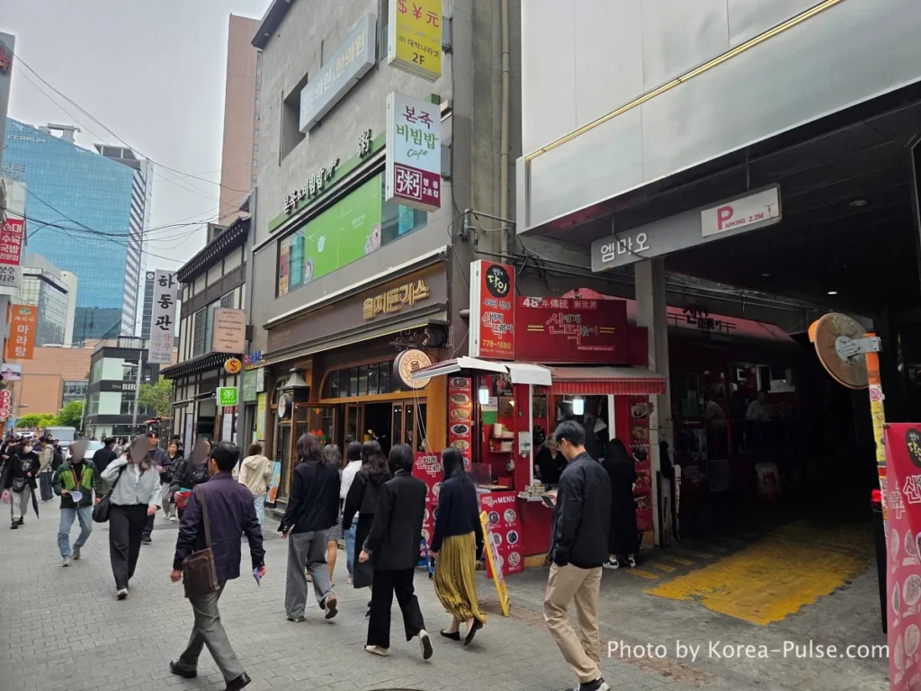 Busy Myeongdong street alley at lunchtime with restaurant signs including Eulji Tonkatsu and Hadongkwan visible, pedestrians walking