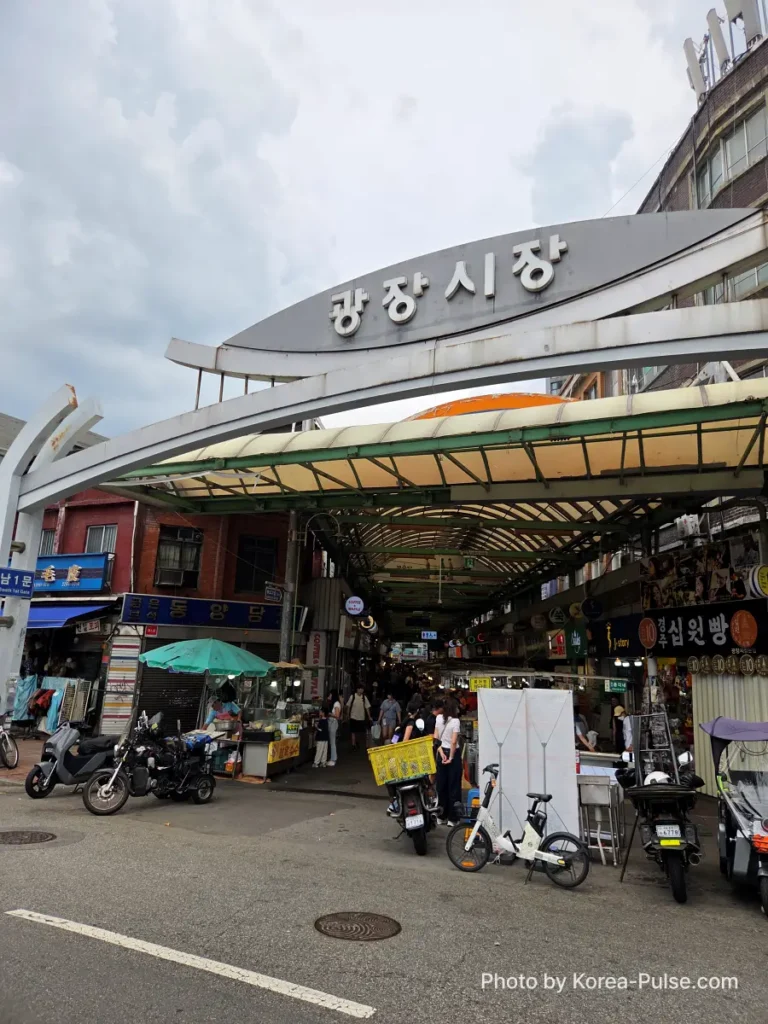 Gwangjang Market Seoul: The Complete Entry Guide — Part 1 6 South Gate 1 of Gwangjang Market Seoul showing the arched 광장시장 sign above a busy covered entrance with motorbikes parked outside