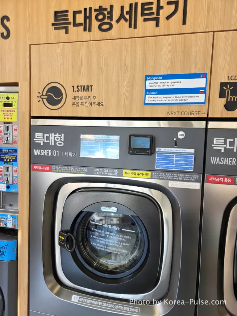 Large industrial washing machine at Easy Wash Korean coin laundry showing front panel with touchscreen course selection and English price labels on coin slot