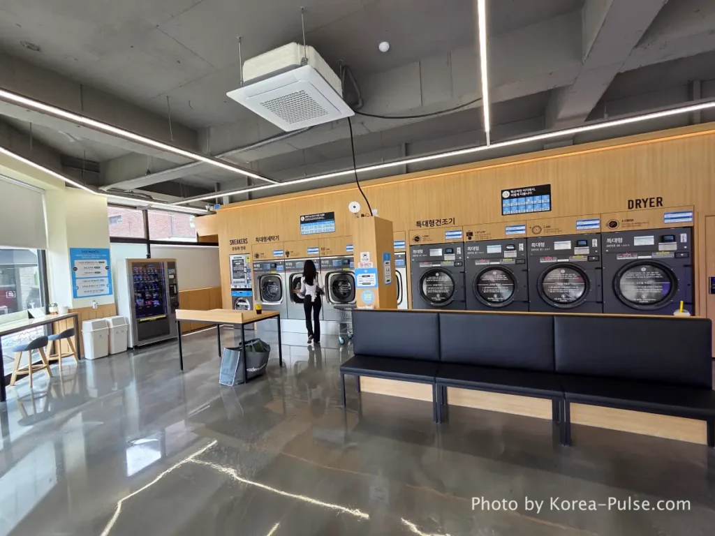 Interior left side of Easy Wash Korean coin laundromat showing rows of large commercial washing machines with cushioned seating along the wall
