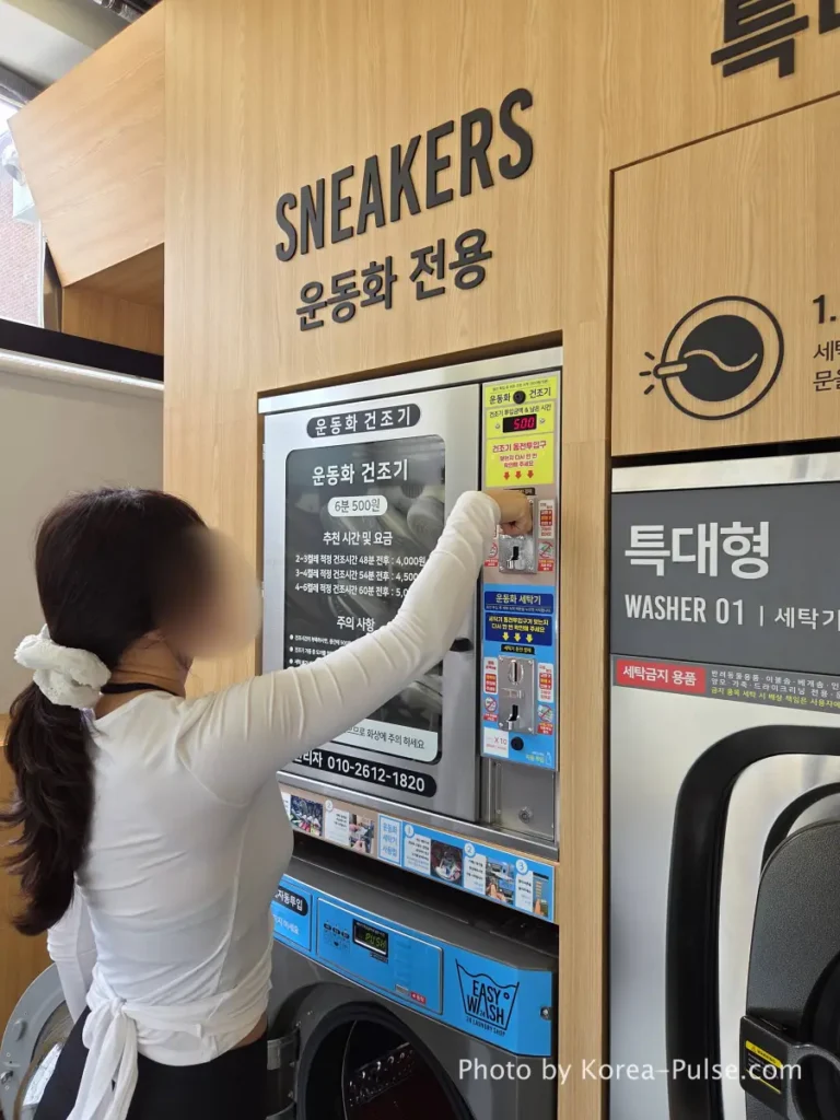 Woman inserting coins into sneaker dryer upper unit at Easy Wash Korean coin laundry with large commercial dryers visible beside sneaker station
