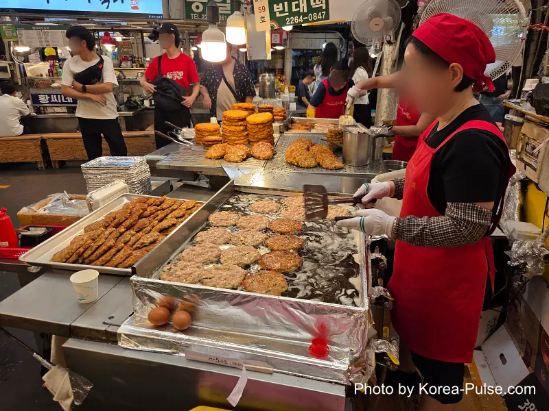 Gwangjang Market bindaetteok vendor in red apron frying mung bean pancakes on griddle Seoul