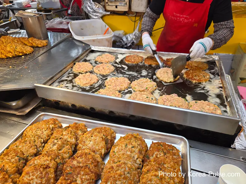 Close-up of golden-brown bindaetteok mung bean pancakes sizzling on griddle Gwangjang Market food alley