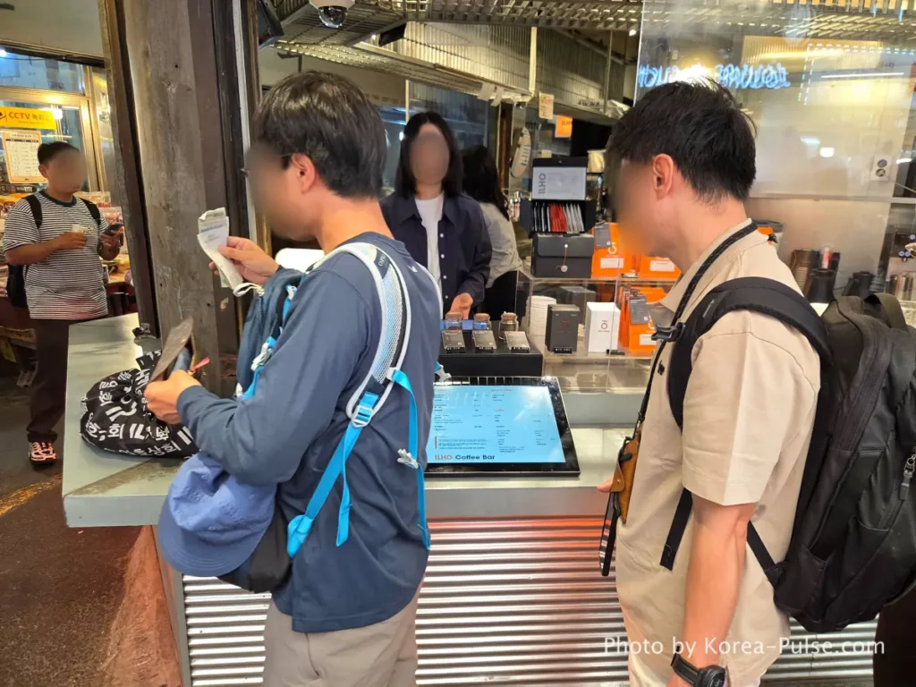 Ilho Coffee Bar Gwangjang Market customer reading menu tablet while another moves to pickup counter