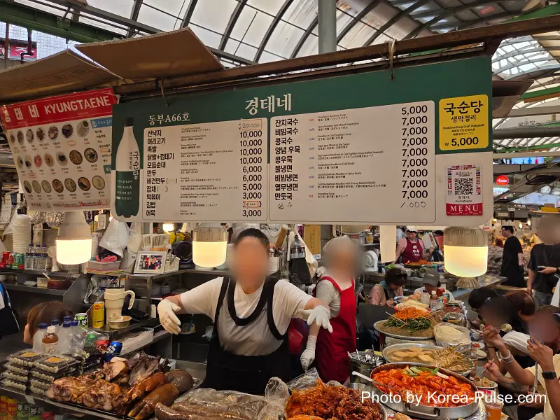 Gwangjang Market food alley vendor serving multiple Korean dishes to international visitors at bench seating
