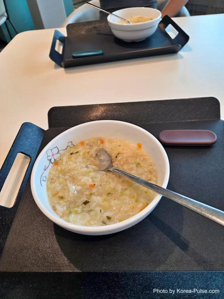 A bowl of warm Korean rice porridge (juk) with a spoon, served on a tray table